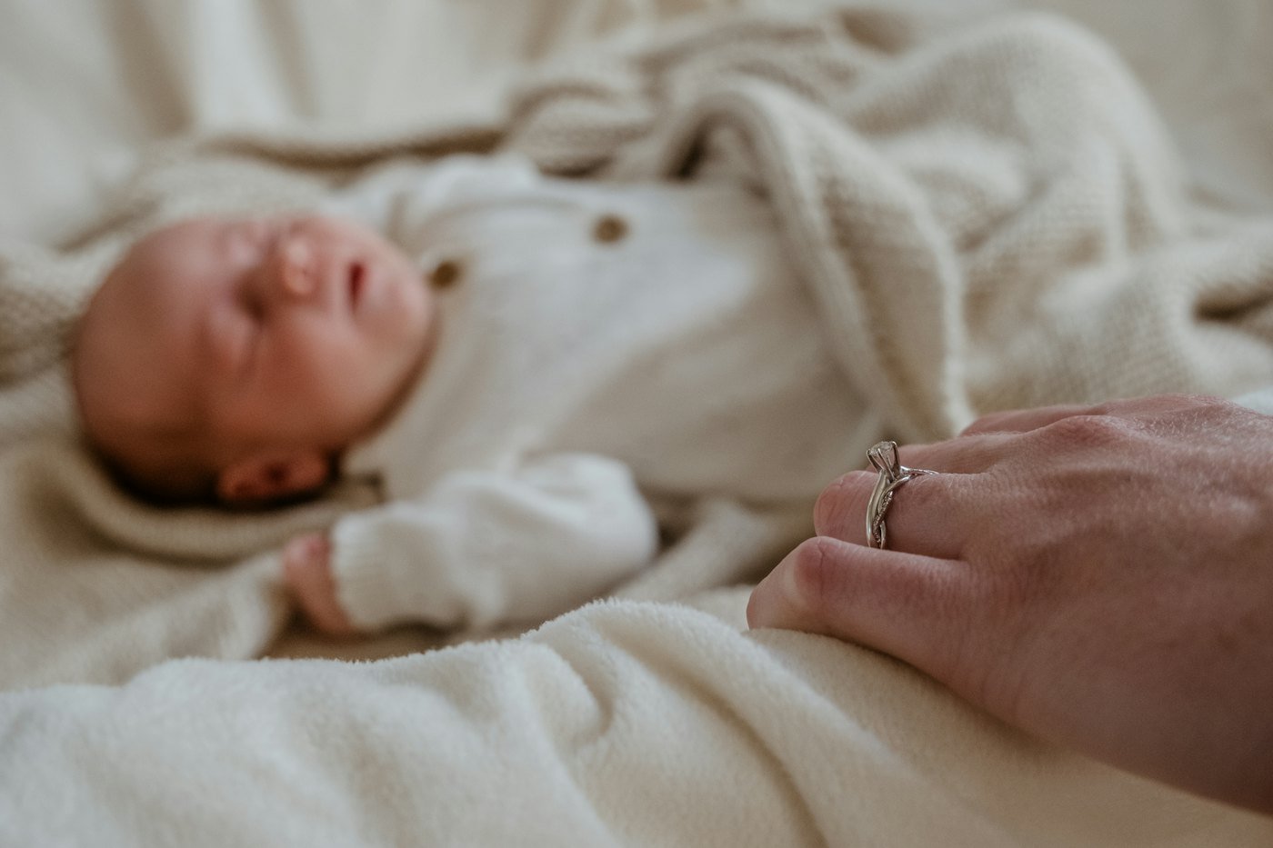 A newborn asleep beside a parent’s hand in soft cream sheets