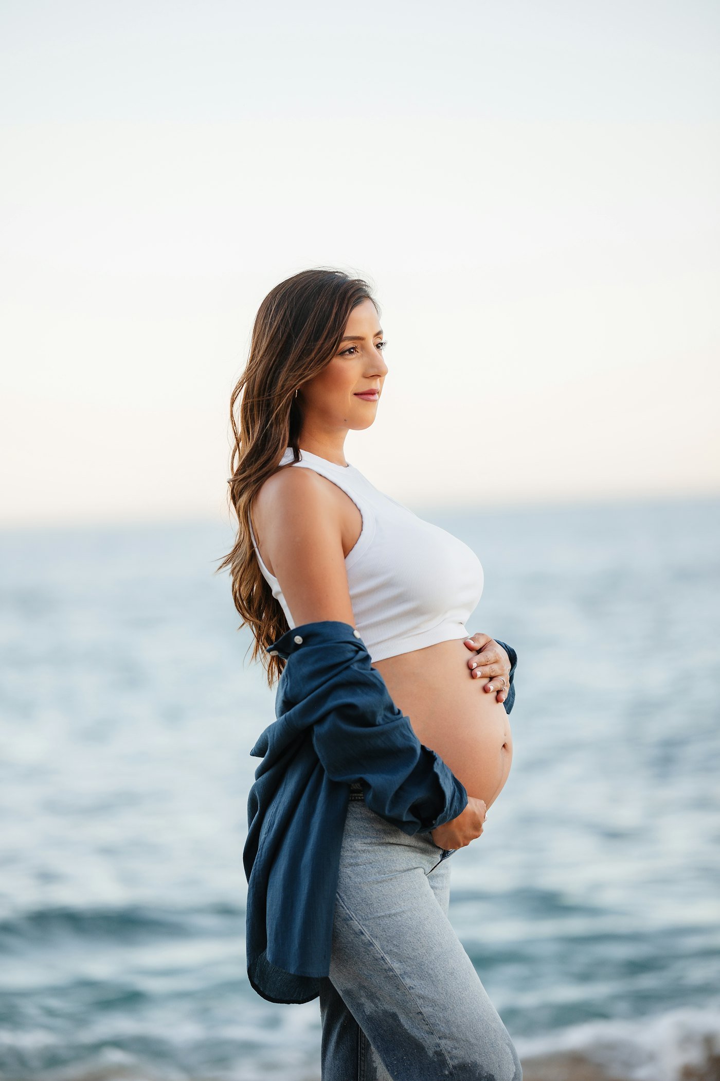 A pregnant woman standing by the sea at golden hour, holding her bump