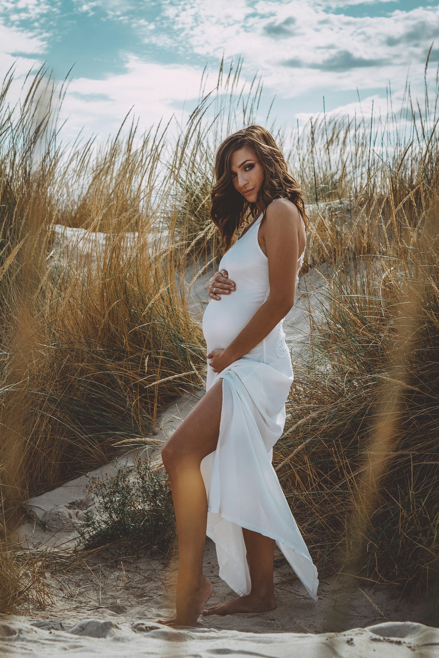 A pregnant woman in a white dress walking through dune grass