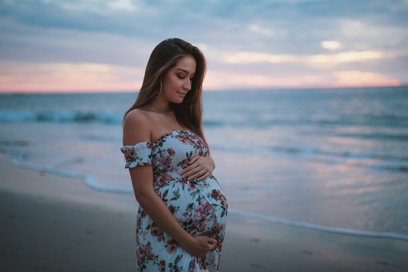 A pregnant woman in a floral dress at the water at dusk